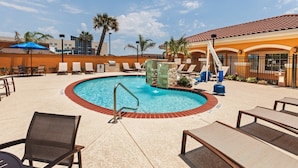 A pool area with lounge chairs, a pool with a red border, and a building with a tiled roof in the background.