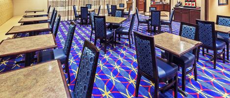 A hotel dining area with multiple tables and chairs, a patterned carpet, and a view of the kitchen in the background.