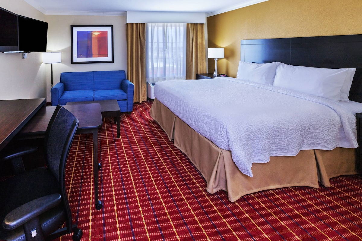 A hotel room with a large bed, a blue sofa, a desk with a chair, a flat-screen TV, and a window with curtains.