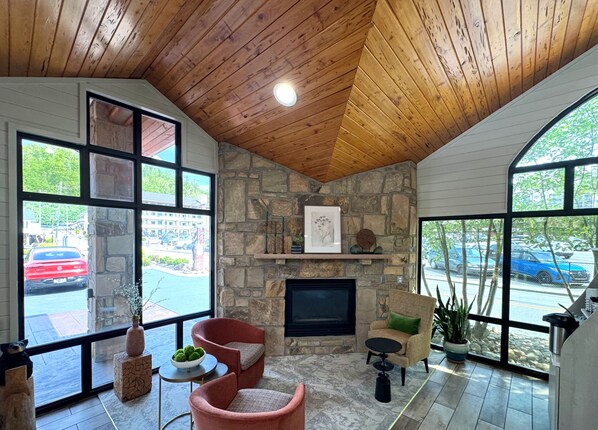 A cozy room with a stone fireplace, large windows, and a wooden ceiling.