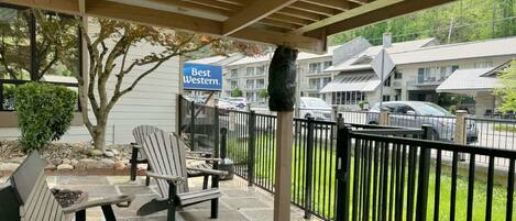 A covered outdoor seating area with Adirondack chairs, a stone-tiled floor, a black metal fence, and a sign for 'Best Western.'