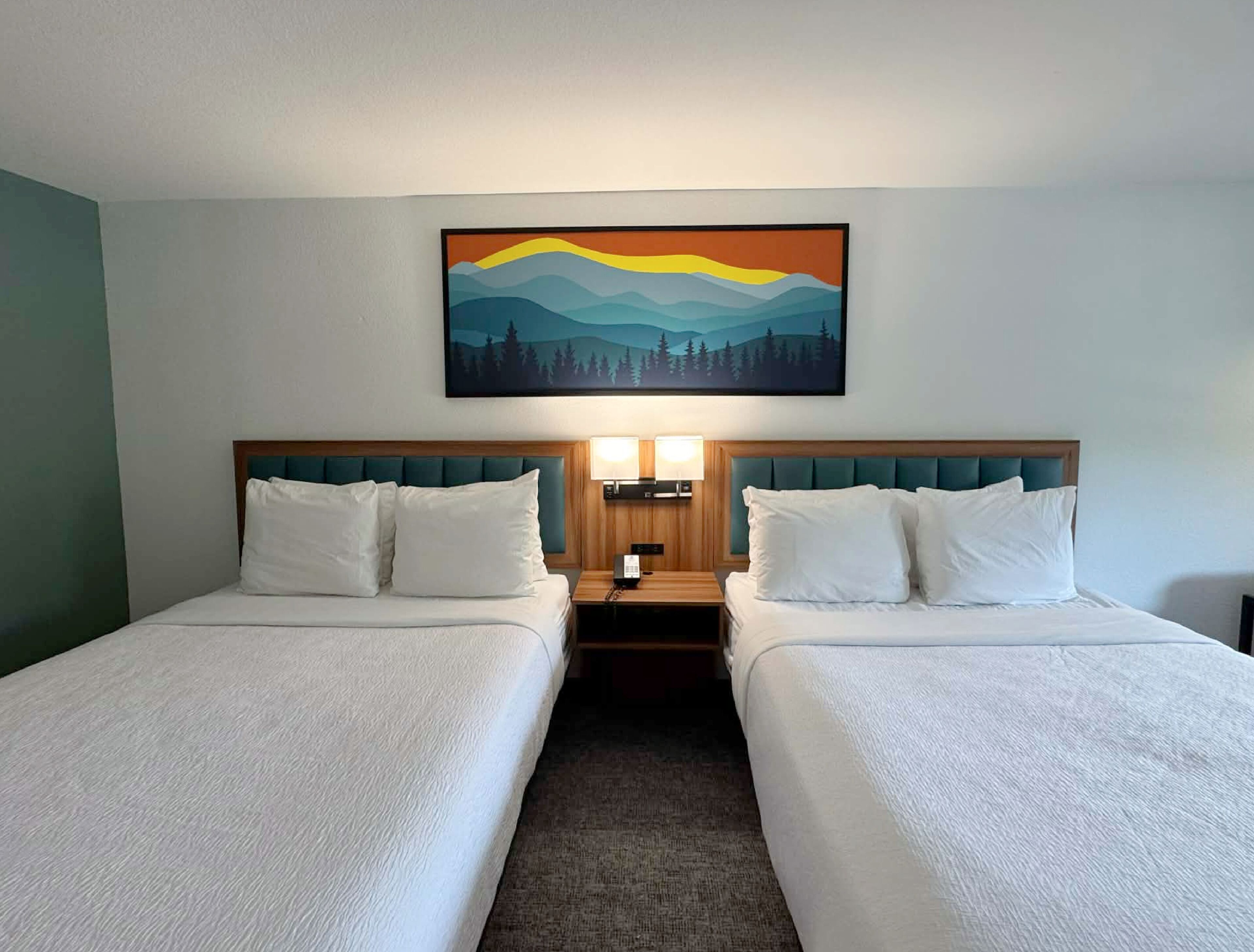 Habitación de hotel con dos camas, un cuadro de un paisaje de montaña y una mesita de noche con una lámpara.
