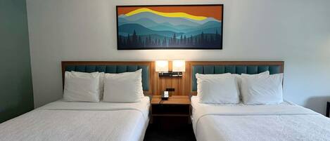 Habitación de hotel con dos camas, un cuadro de un paisaje de montaña y una mesita de noche con una lámpara.