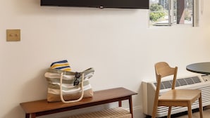 Habitación con una televisión de pantalla plana montada en la pared, un banco, una silla, una mesa y una ventana con vista a zonas verdes.