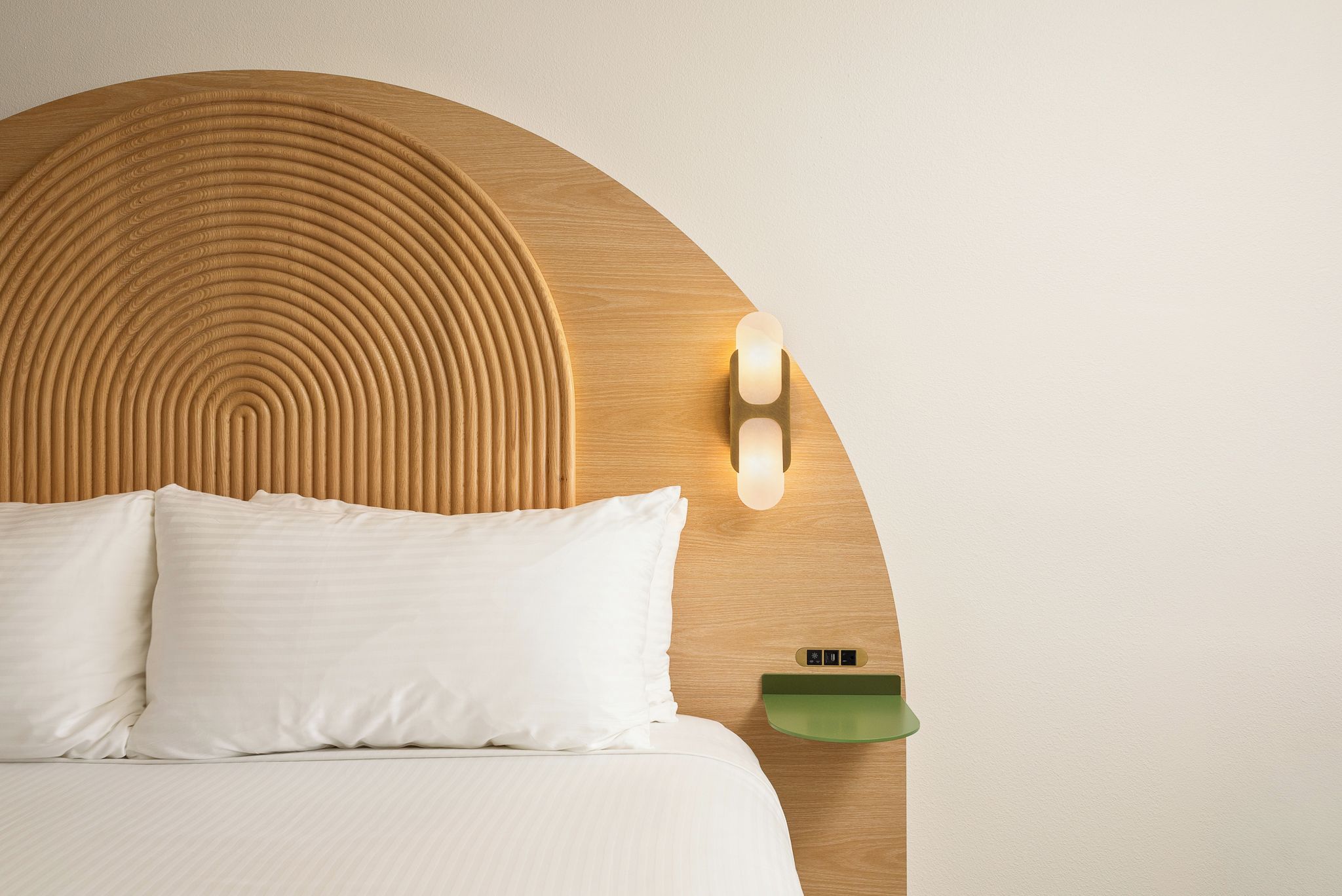 Una cama con cabecera de madera, dos almohadas, una mesita de noche con una bandeja verde y una lámpara fijada a la pared.