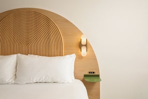 Una cama con cabecera de madera, dos almohadas, una mesita de noche con una bandeja verde y una lámpara fijada a la pared.