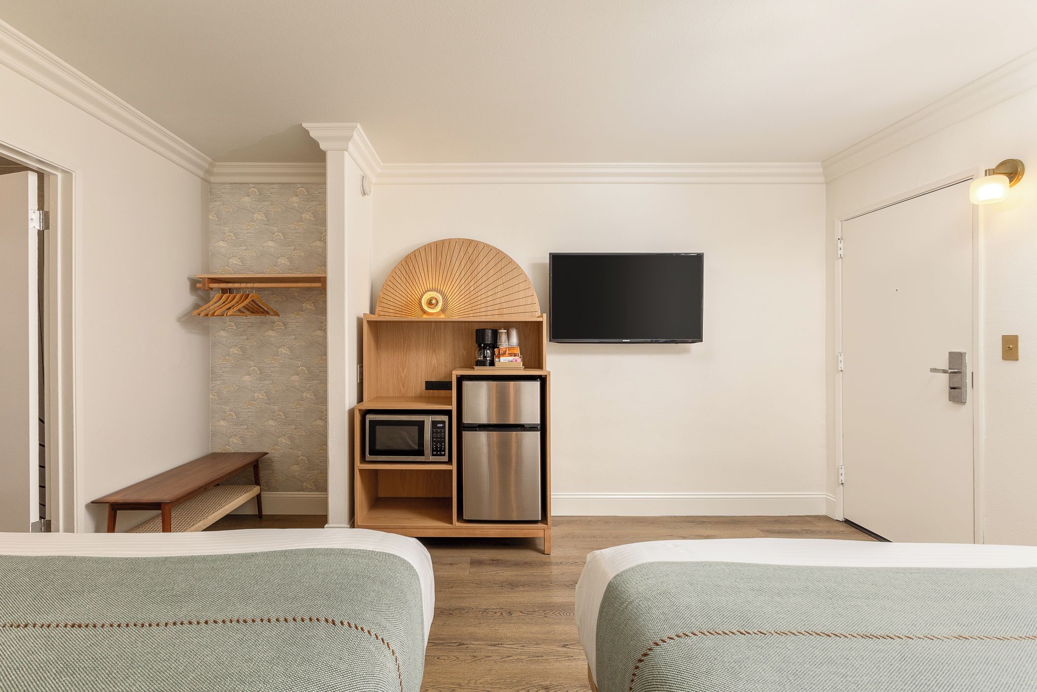 Una habitación de hotel con dos camas, una kitchenette pequeña con microondas y refrigerador, un televisor montado en la pared y un banco.