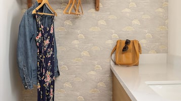 A bathroom with a wooden shelf holding a hat and a handbag, a floral dress, and a pair of sandals.