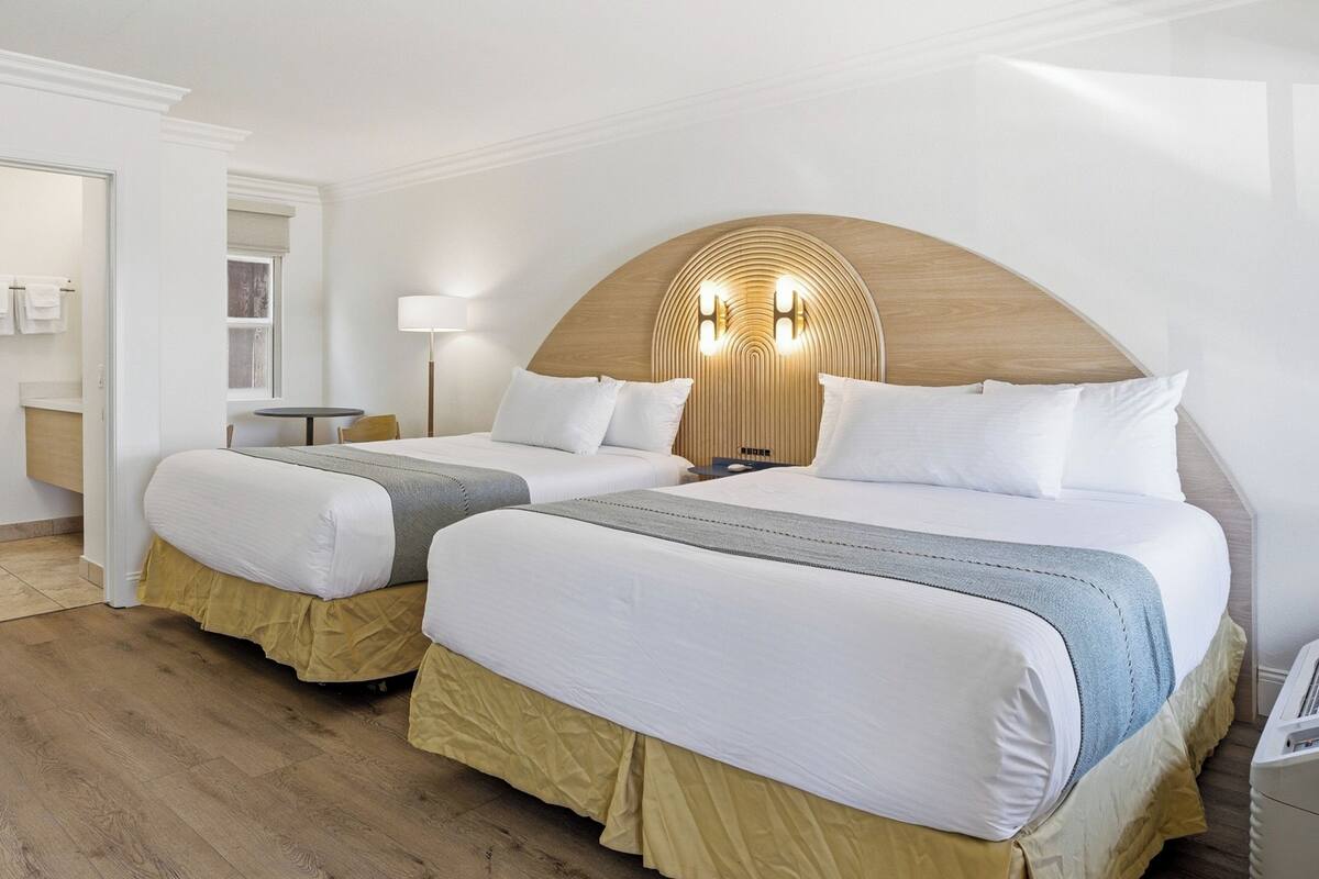A hotel room with two beds, a wooden headboard with lights, a small table, and a mirror.