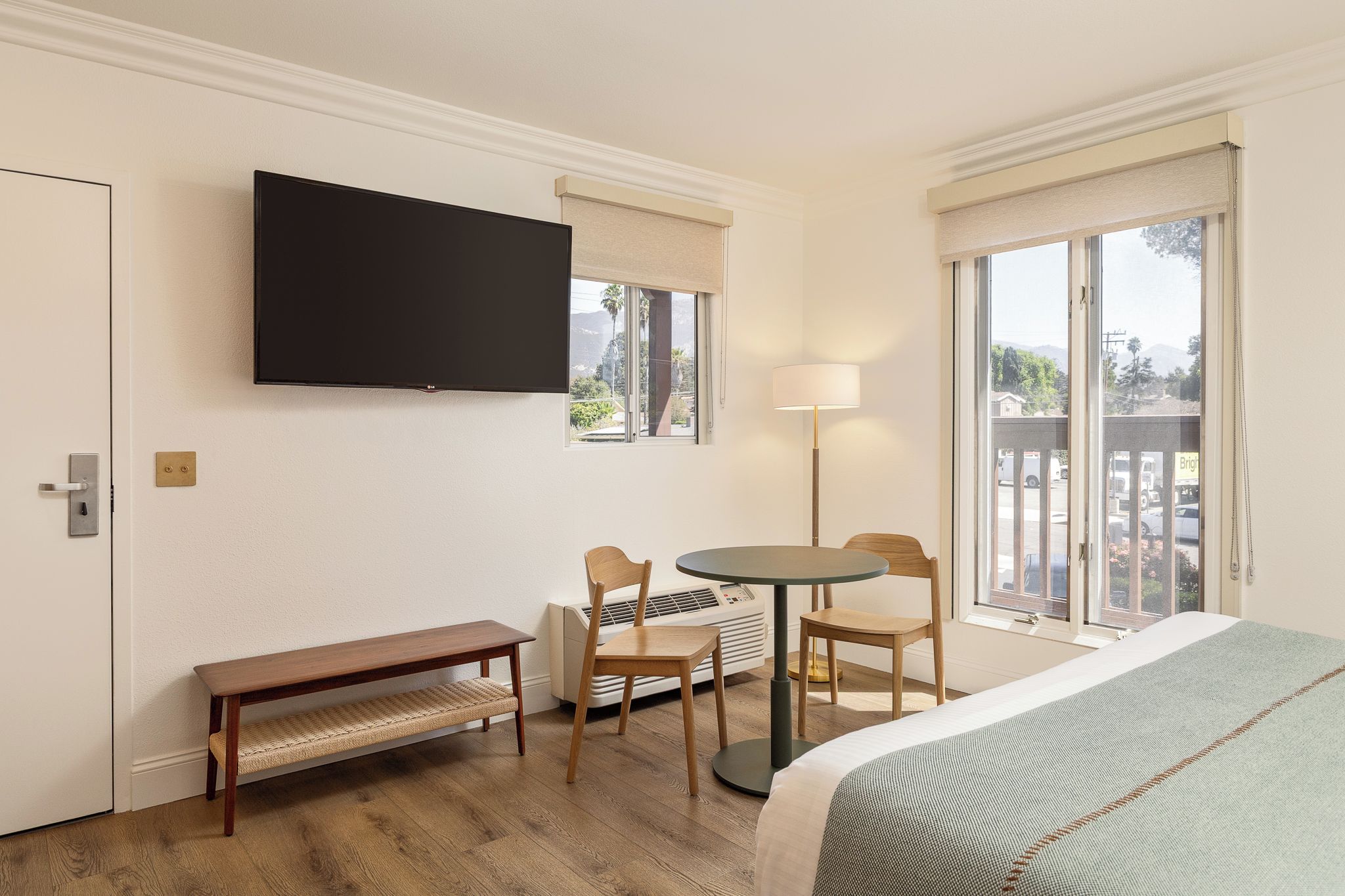 A hotel room with a bed, a TV mounted on the wall, a desk with chairs, a lamp, and a view of the outdoors through the windows.