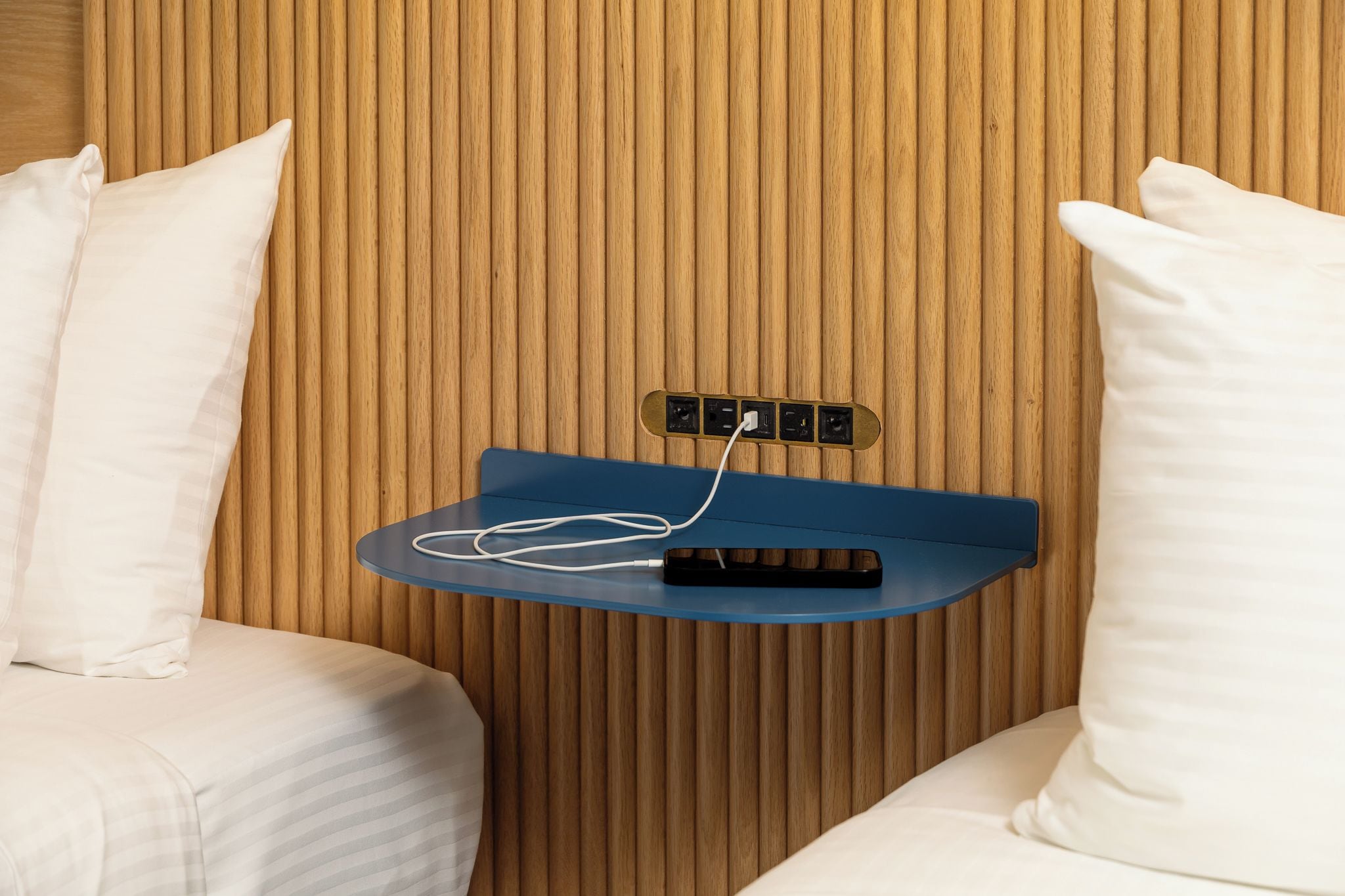 Quarto de hotel com parede de painel de madeira, mesa de cabeceira azul com controle remoto e travesseiros brancos.