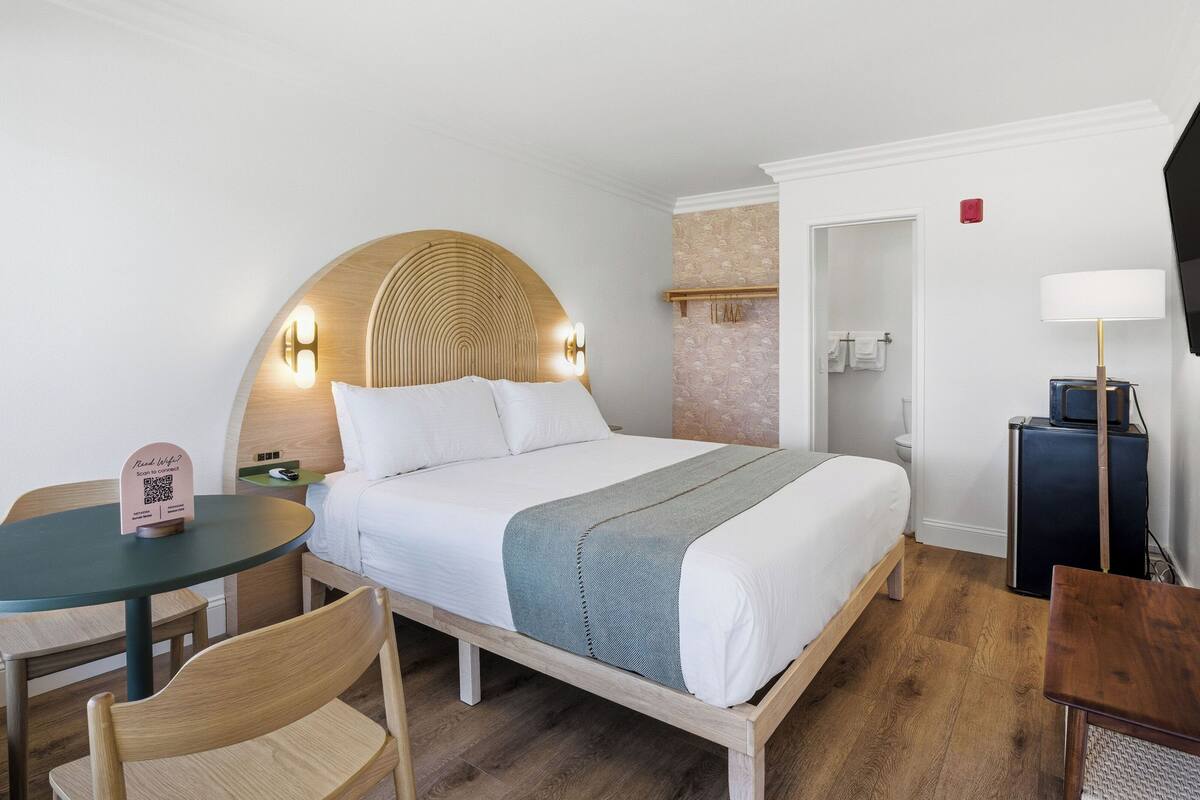 A neatly arranged hotel room with a bed, a small table, a chair, a television, and a door leading to the bathroom.