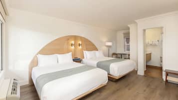 A hotel room with two beds, a wooden headboard, bedside tables, a desk, and a bathroom visible through an open door.