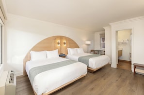 A hotel room with two beds, a wooden headboard, bedside tables, a desk, and a bathroom visible through an open door. - Solei House (Santa Barbara)