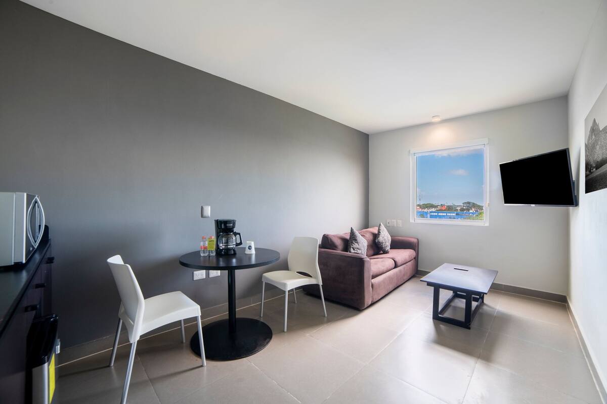Une pièce moderne avec un mur gris, un canapé rose, une chaise blanche, une petite table ronde avec une machine à café, un téléviseur fixé au mur et une fenêtre donnant sur la ville.