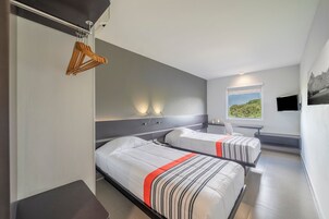 Habitación de hotel moderna con dos camas, un perchero de madera, una mesita de noche, un televisor y una ventana con vistas a zonas verdes.