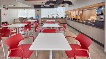 Una cafetería moderna con sillas rojas y mesas blancas, una máquina de café y un dispensador de agua.