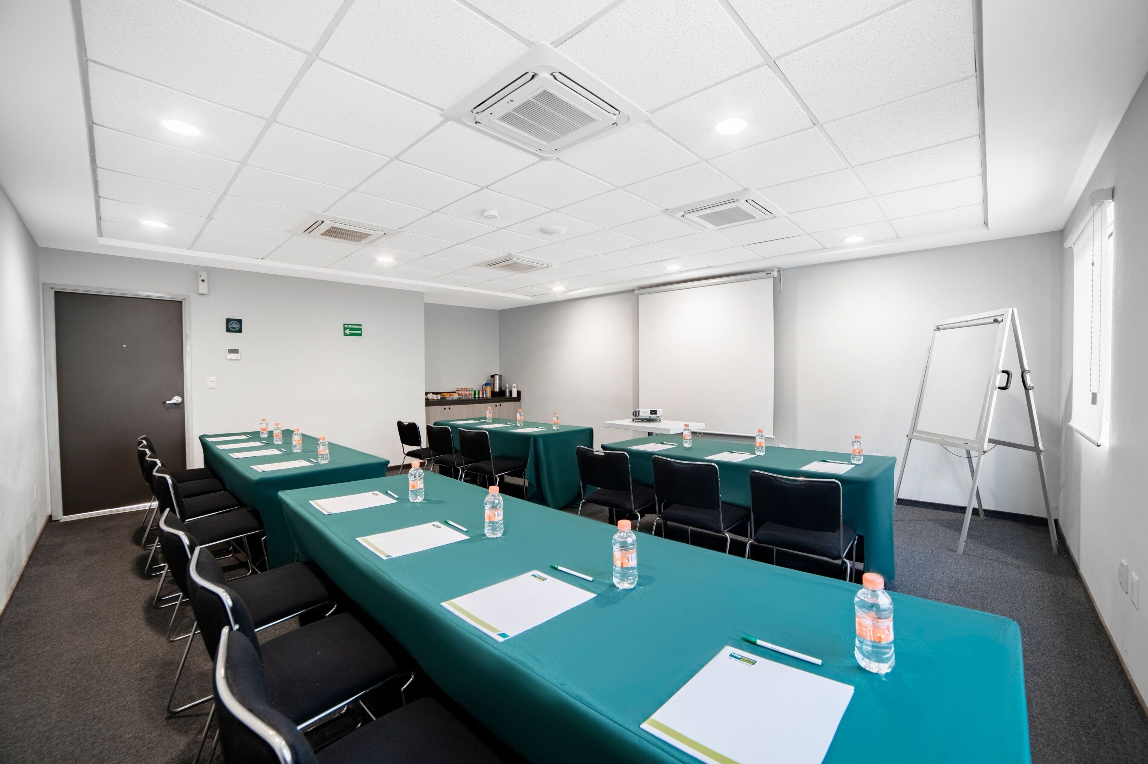Une salle de conférence équipée de longues tables, de chaises, de bouteilles d’eau et d’un tableau blanc.