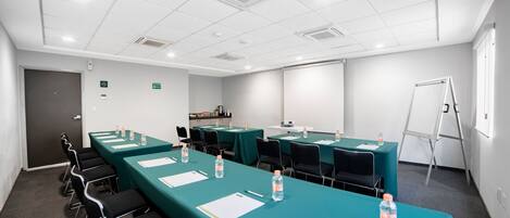 Une salle de conférence équipée de longues tables, de chaises, de bouteilles d’eau et d’un tableau blanc.