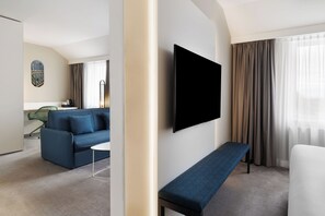 Una habitación de hotel moderna con un televisor de pantalla plana montado en la pared, un sofá azul, una pequeña mesa de centro, un banco y vistas de la ciudad a través de amplias ventanas con cortinas.