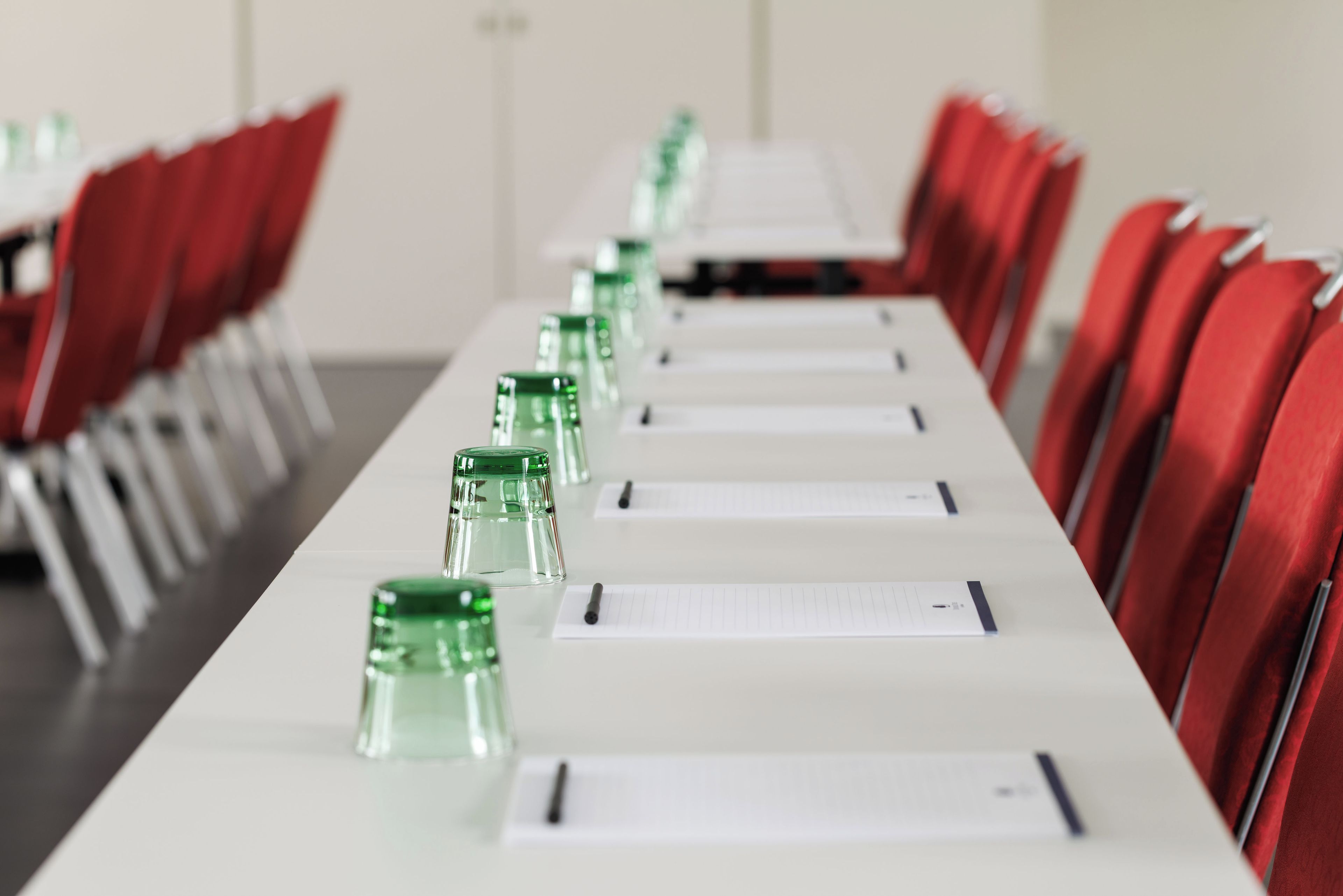 Sala de conferência com uma mesa comprida, cadeiras vermelhas e garrafas de água de vidro verde.