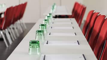 Sala de conferência com uma mesa comprida, cadeiras vermelhas e garrafas de água de vidro verde.