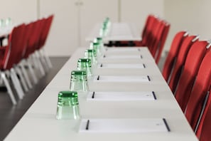 Un salón de conferencias con una larga mesa, sillas rojas y botellas de agua de vidrio verde.