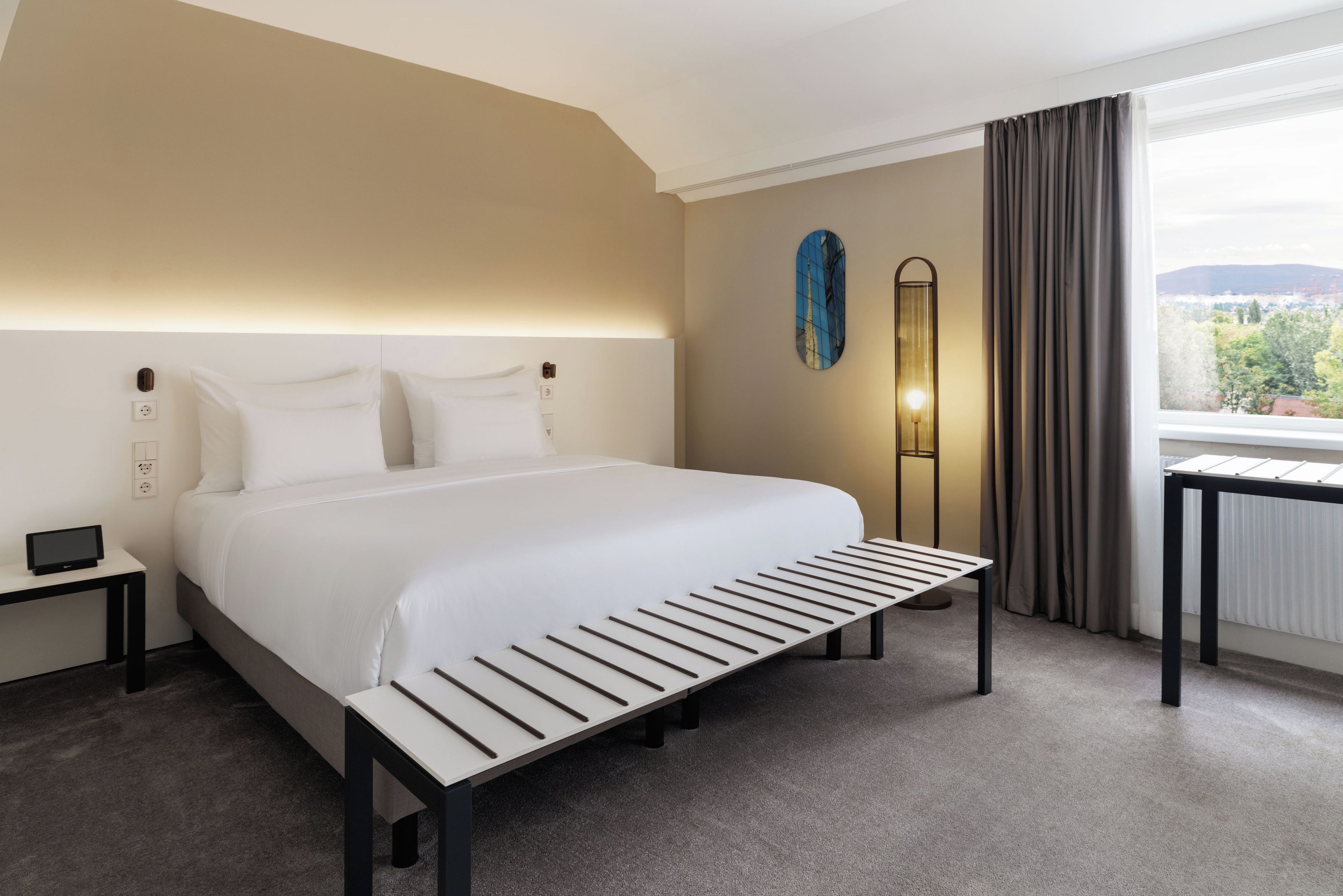 Een hotelkamer met een groot bed, nachtkastjes, een bedlampje, uitzicht op het omliggende landschap en een raam met gordijnen.