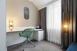 Una habitación de hotel con escritorio, una silla verde, una lámpara de pie, una luz empotrada en la pared y una ventana con cortinas.
