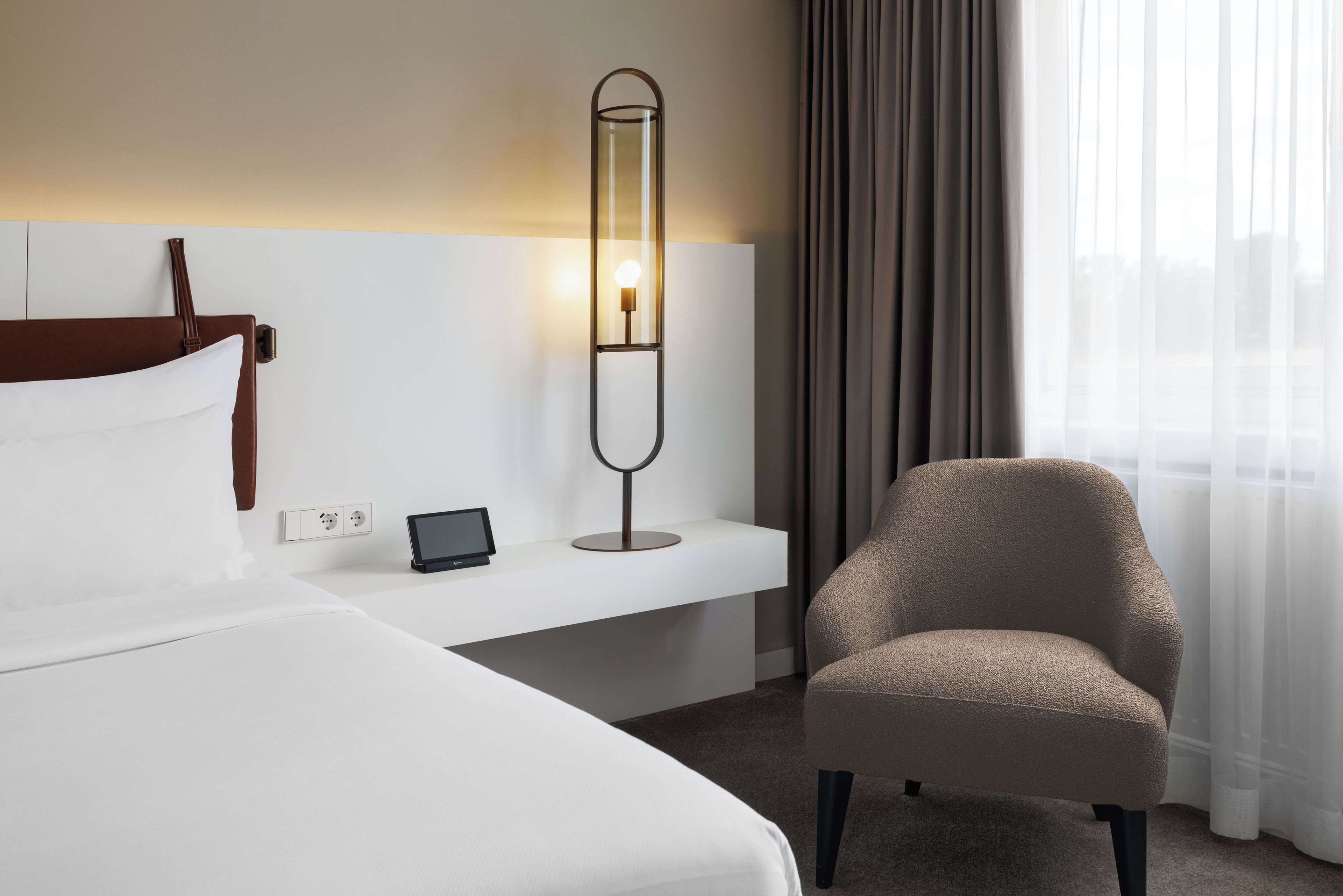 Hotellihuone, jossa on sänky, yöpöytä lampun kanssa, tuoli ja näkymä kaupunkiin ohuiden verhojen läpi.