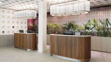 A hotel reception area with a modern design, featuring a wooden reception desk, a large chandelier, and a patterned ceiling.