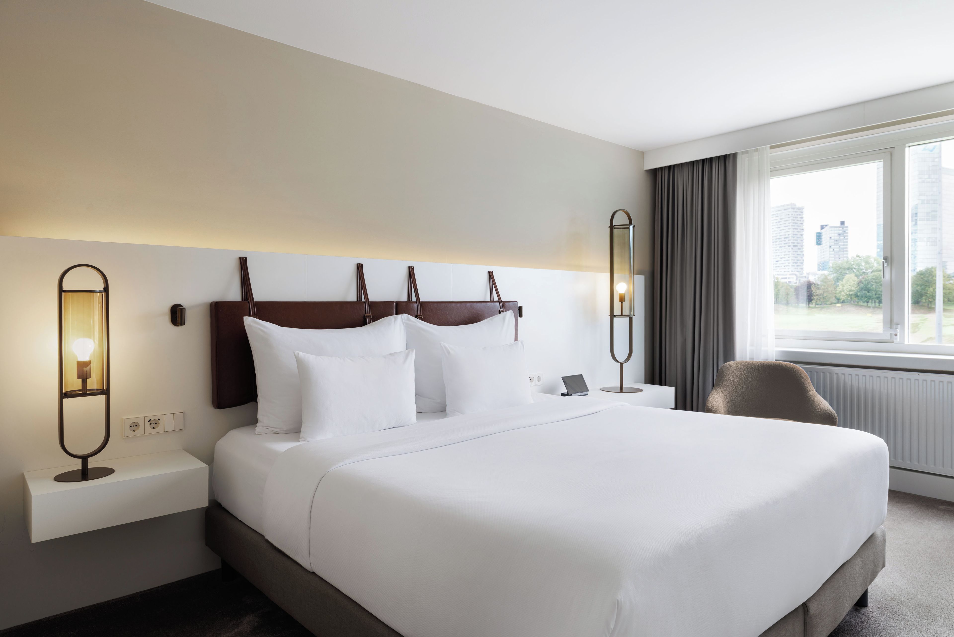 Habitación de hotel con una cama grande, dos mesitas de noche, una silla y vistas a la ciudad desde la ventana.