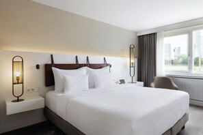 A hotel room with a large bed, two bedside tables, a chair, and a view of the city through the window. - DoubleTree by Hilton Vienna Bosei (Vienna)