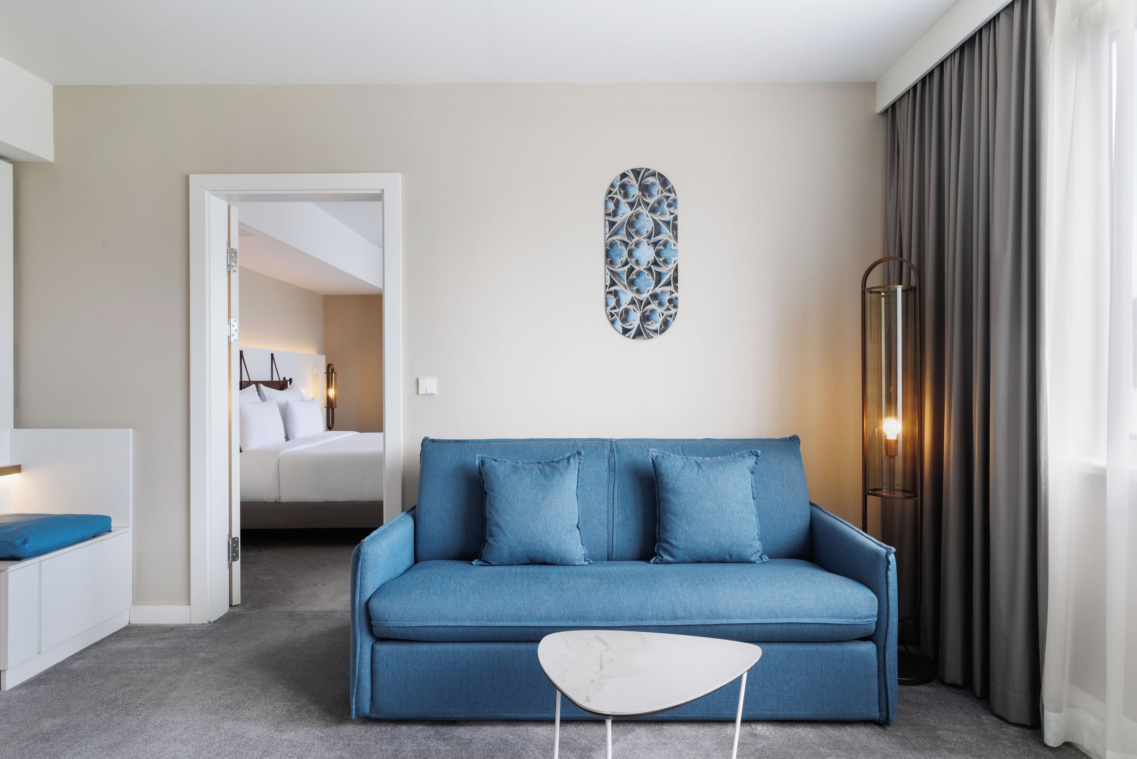 A modern hotel room with a blue sofa, a white side table, a decorative wall piece, and a large window with curtains.