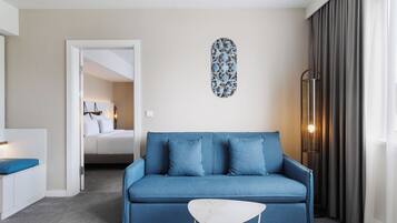A modern hotel room with a blue sofa, a white side table, a decorative wall piece, and a large window with curtains.