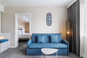 Habitación de hotel moderna con un sofá azul, una mesita auxiliar blanca, un adorno de pared y un ventanal con cortinas.