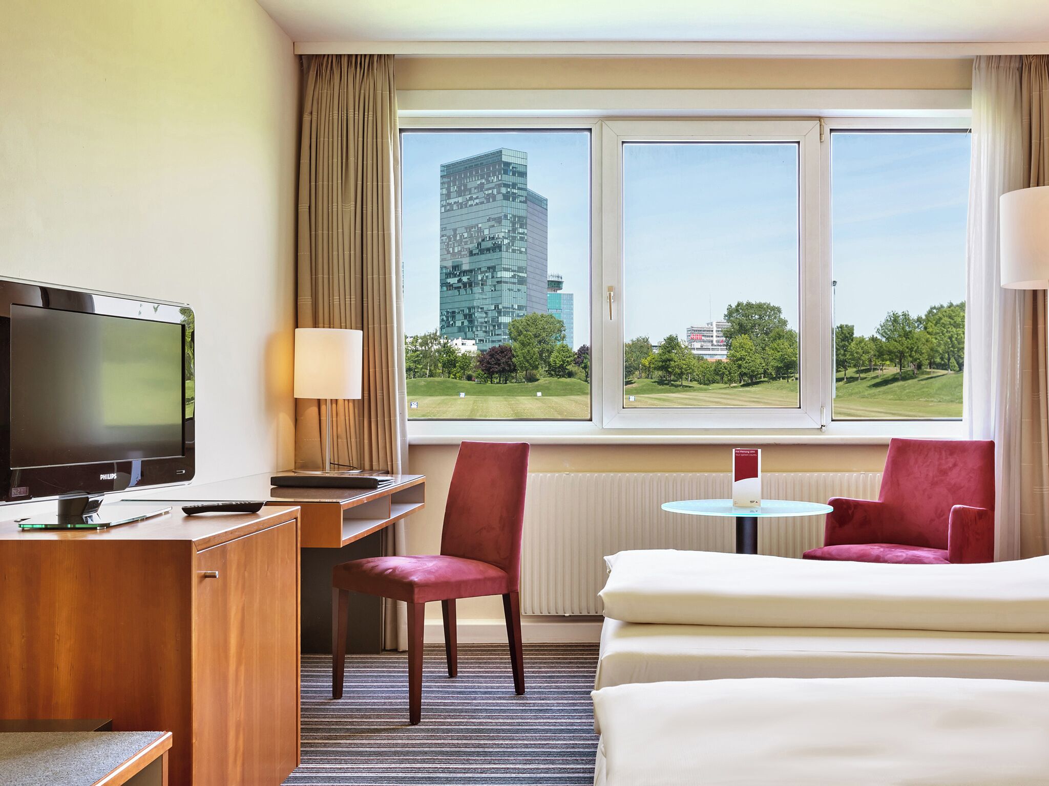 Habitación de hotel con cama, escritorio con televisor, silla, una mesita y vistas a un edificio y parque.
