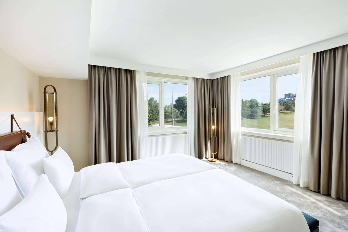 A hotel room with a large bed, a nightstand, a lamp, and a view of the outdoors through large windows.