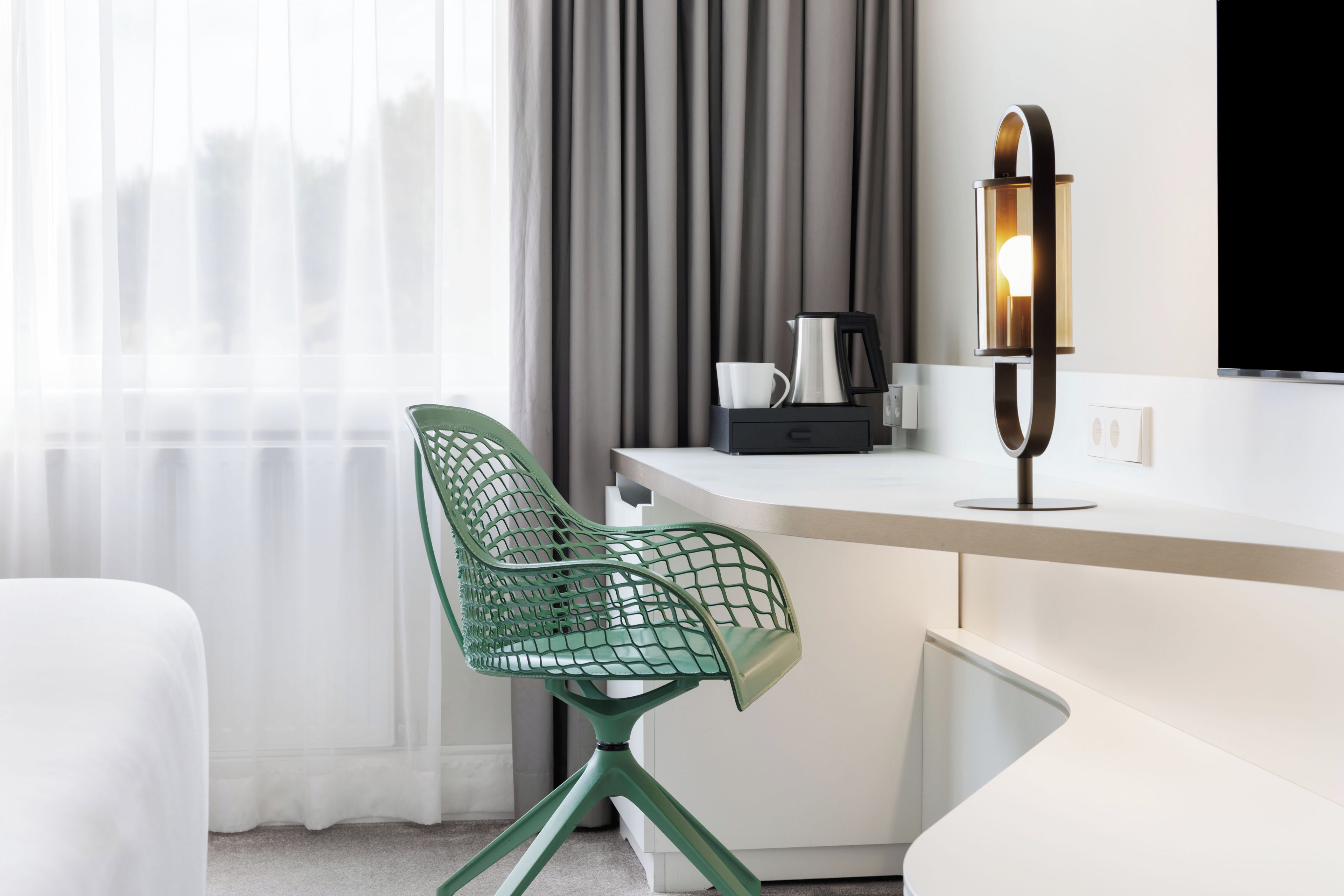 A modern hotel room with a green mesh chair, a white desk, a coffee maker, and a wall-mounted lamp.