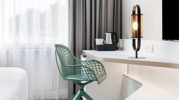 A modern hotel room with a green mesh chair, a white desk, a coffee maker, and a wall-mounted lamp.
