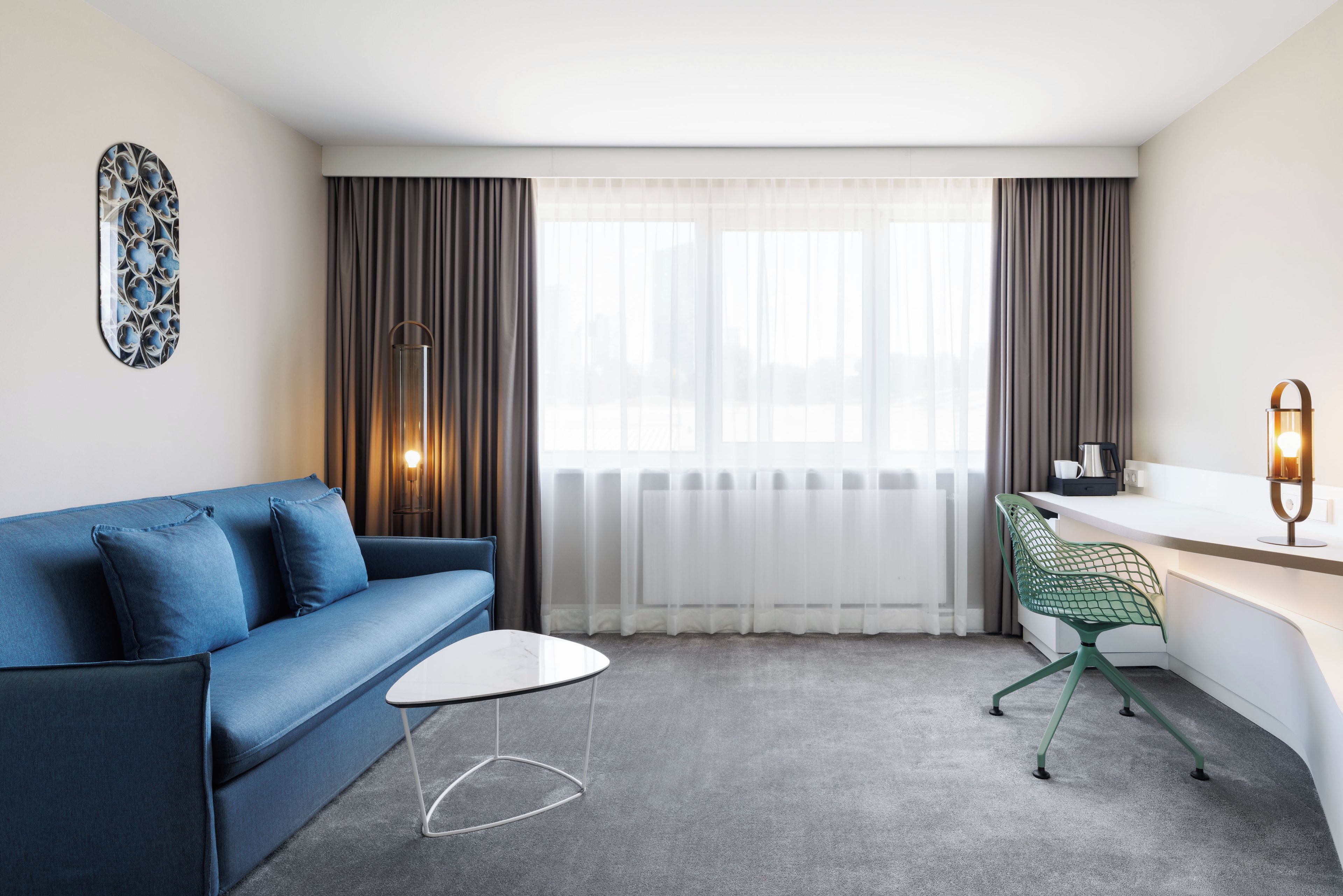 Ein modernes Hotelzimmer mit einem blauen Sofa, einem kleinen weißen Tisch, einem grünen Sessel, einem Schreibtisch und einem großen Fenster mit Vorhängen.