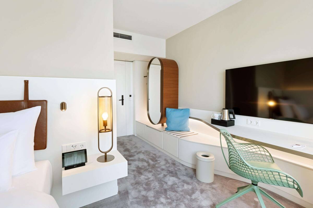 A modern hotel room with a bed, a desk with a chair, a large mirror, a TV, and a small trash bin.