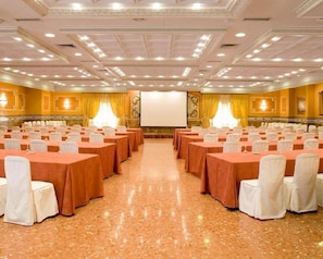 Une salle de conférence avec des rangées de tables, des chaises blanches et un grand écran à l’avant.