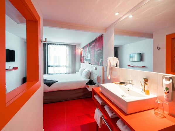 Habitación de hotel con cama, espejo grande, lavamanos y televisor.
