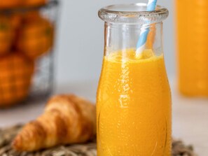 En glasflaska med apelsinjuice och ett blått sugrör, ett croissant och en suddig bakgrund med apelsiner.