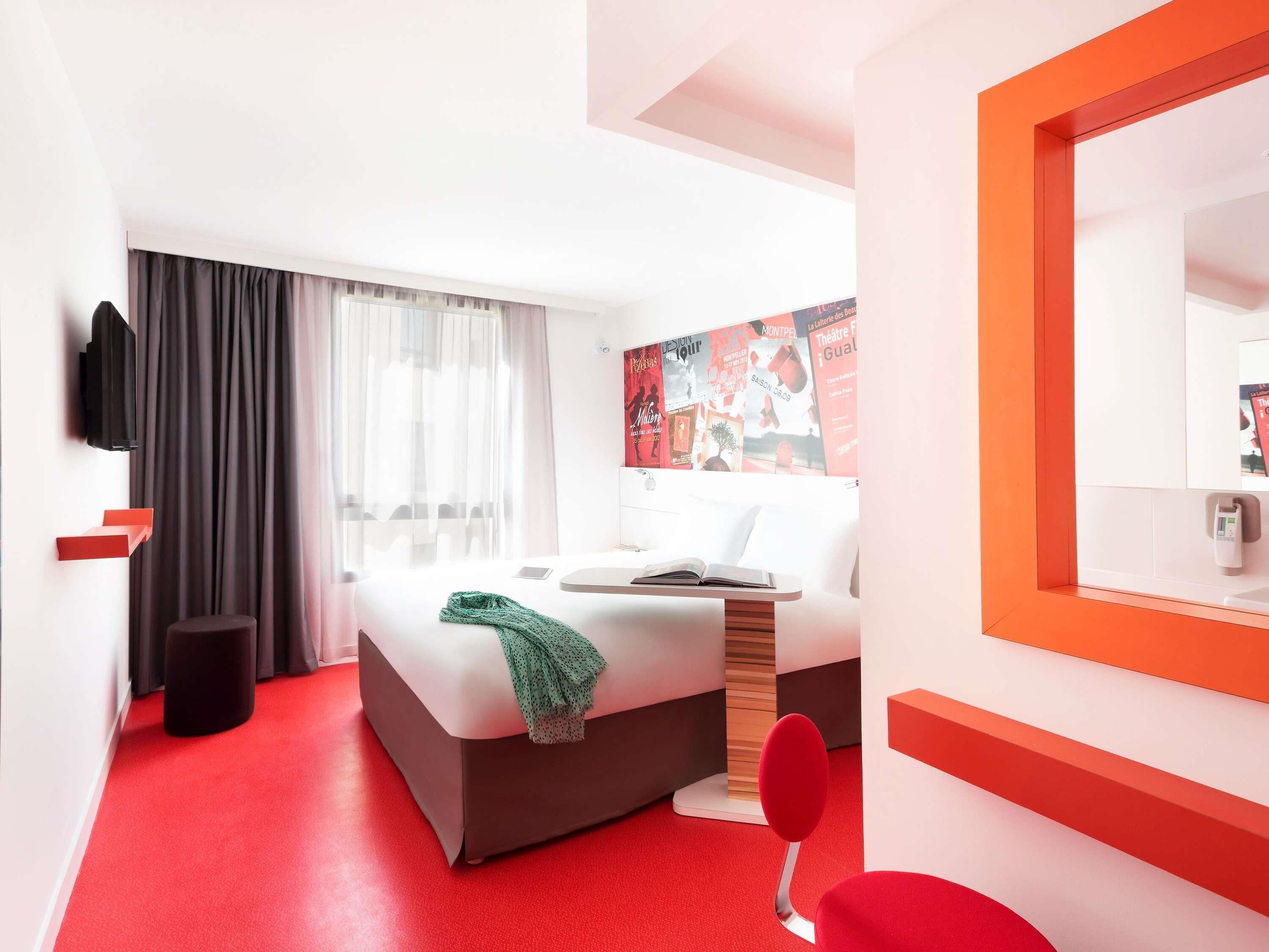Ett modernt hotellrum med en säng, en röd stol, en stor spegel och en röd matta.