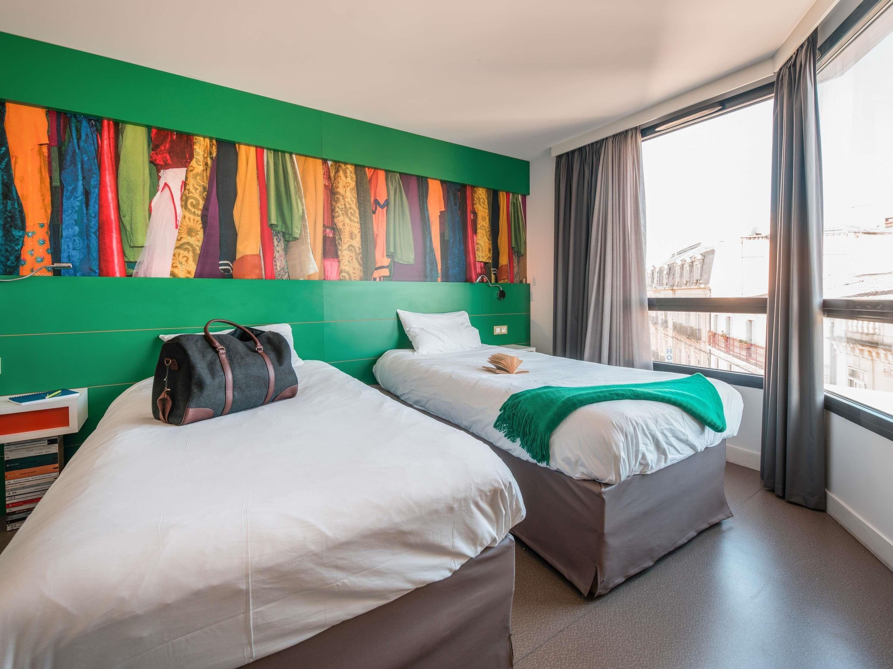 Une chambre d’hôtel avec deux lits, un mur d’accent vert, une fenêtre avec des rideaux et une vue sur les bâtiments environnants.