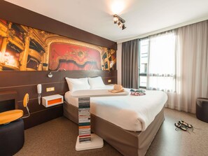 Ein Hotelzimmer mit einem großen Bett, einem Nachttisch, einem Telefon, einem Bücherregal und einem Fenster mit Vorhängen.