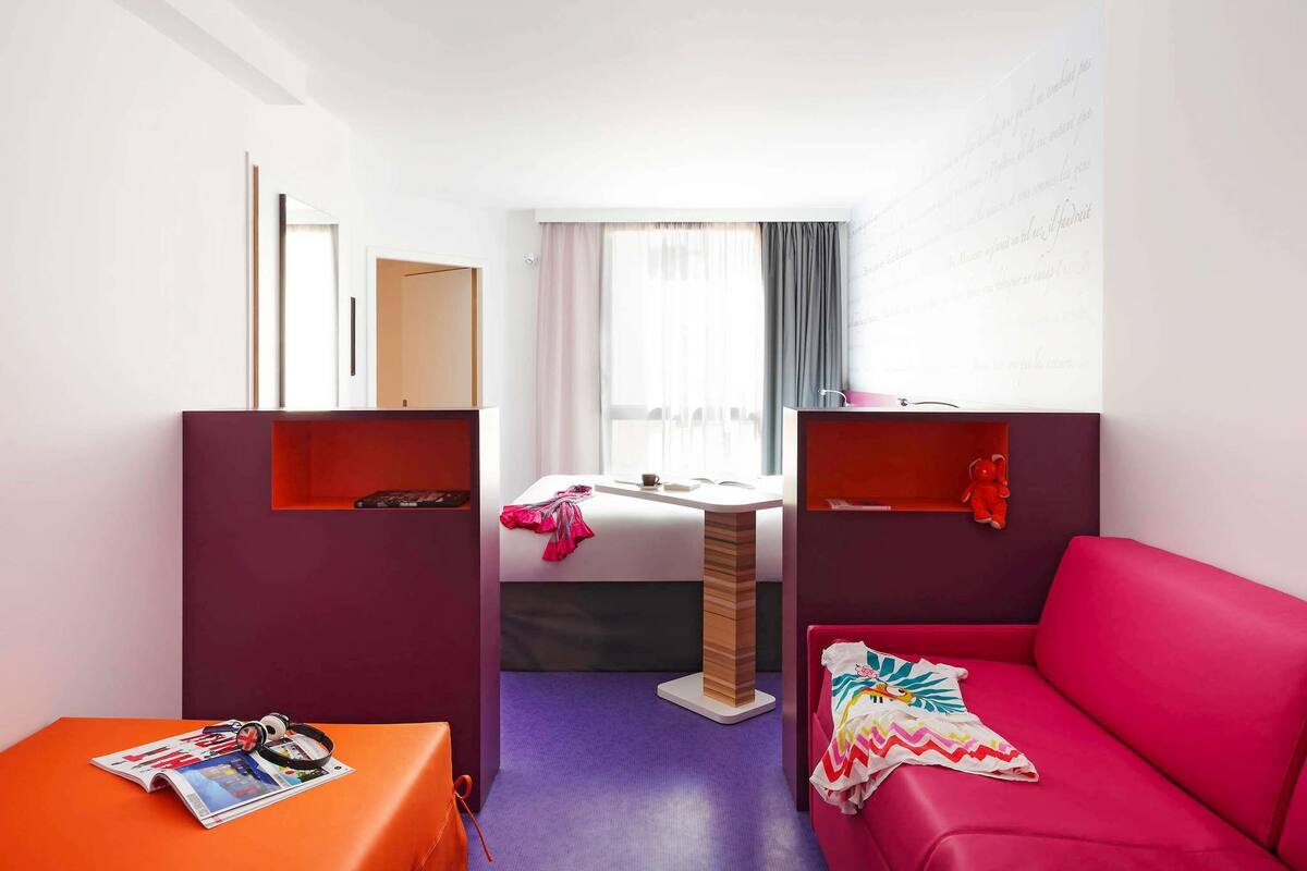 Ein Hotelzimmer mit Bett, Schreibtisch, Stuhl, Sofa und einem Fenster mit Vorhängen.