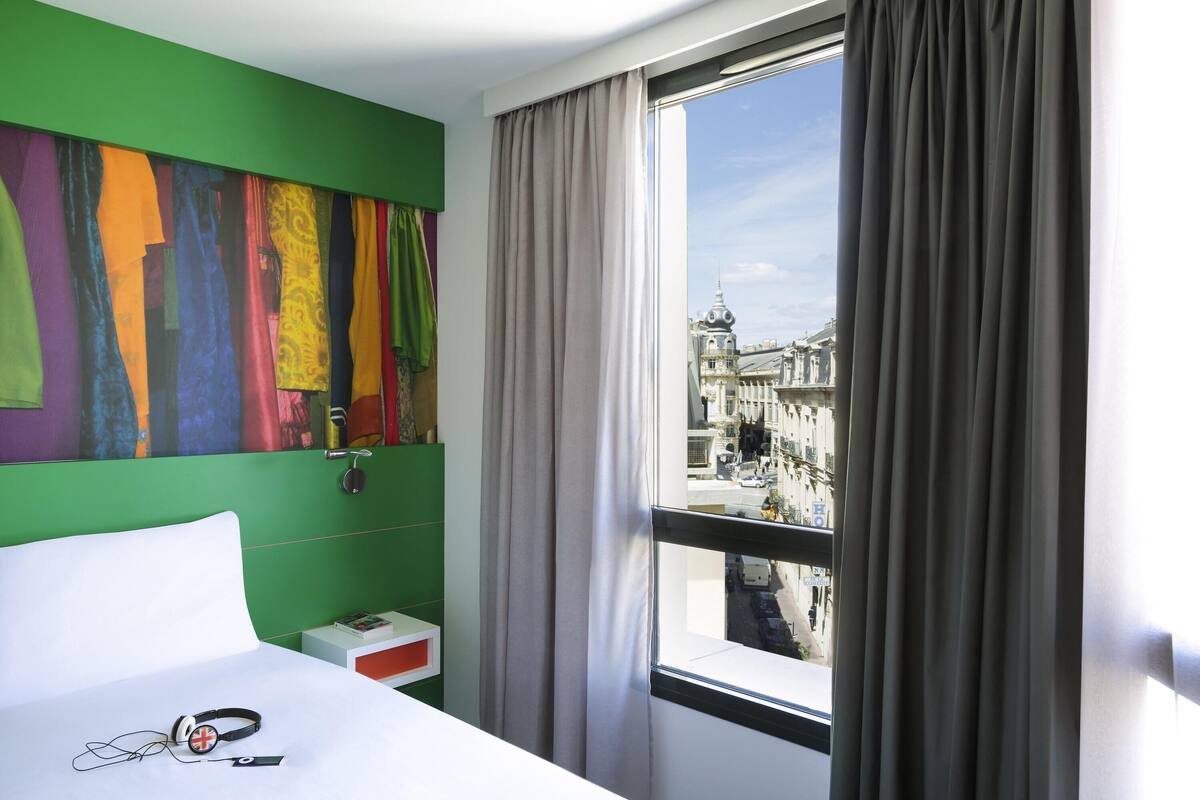 Ein Hotelzimmer mit einem Bett, einer grünen Wand mit farbenfrohen Kunstwerken, einem Fenster mit Vorhängen und Blick auf eine Stadtlandschaft.